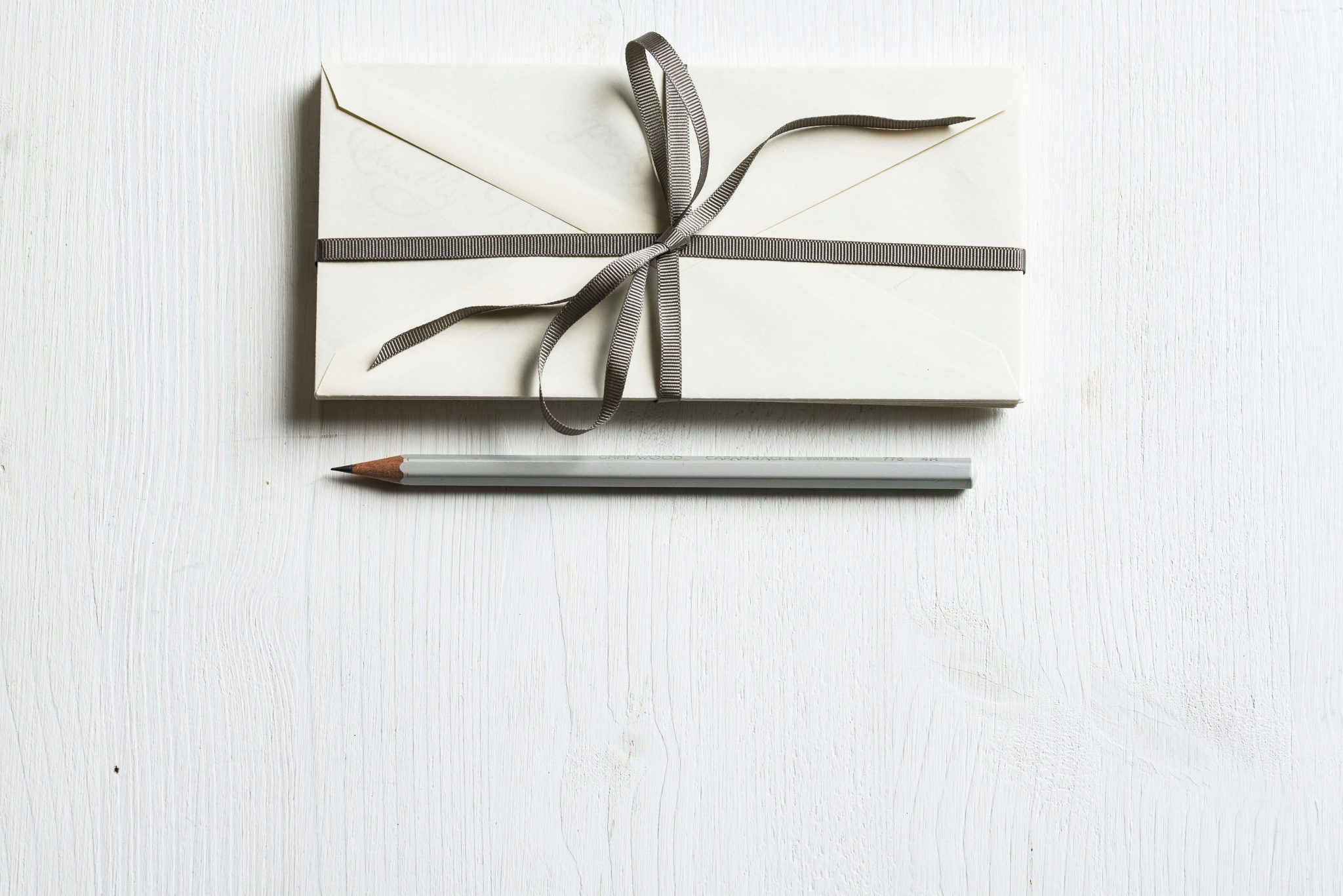 An off-white or cream-colored envelope tied with a dark gray ribbon, lying on a textured white wooden surface. A gray pencil is placed horizontally below the envelope. The image suggests correspondence, a special delivery,